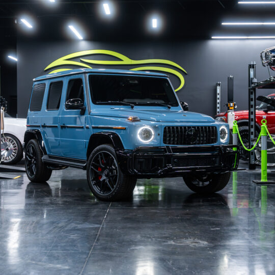 The 2025 Mercedes-Benz G63 AMG in a striking blue color features a robust design with prominent round headlights and black wheels, showcased in a modern automotive display setting.