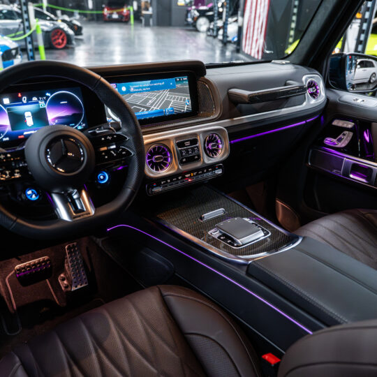 The interior of the 2025 Mercedes-Benz G63 AMG features a modern design with black leather seats, a digital display, and ambient lighting. The dashboard includes user-friendly controls and a stylish steering wheel with integrated buttons.