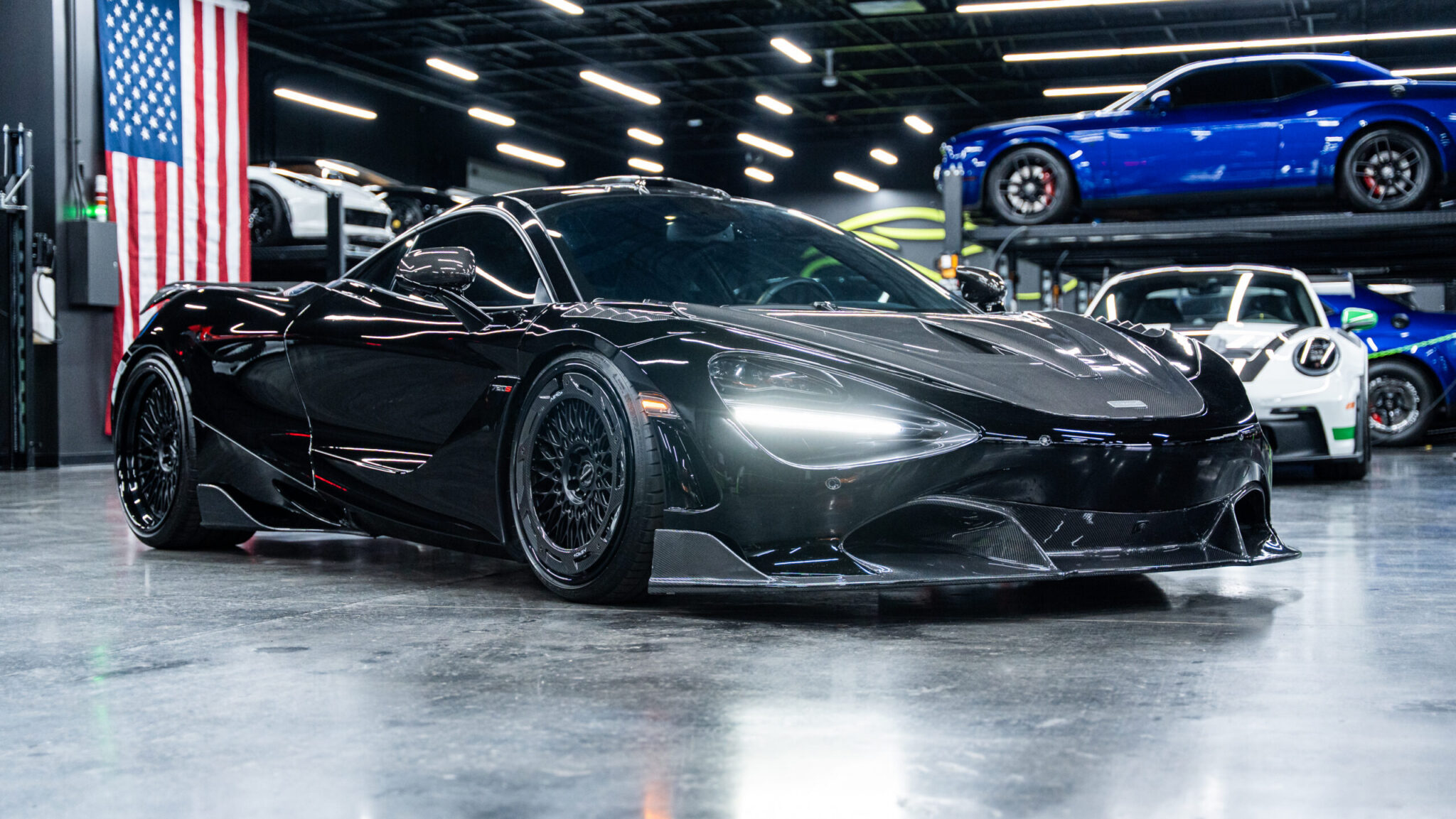 A low-profile black sports car with sleek aerodynamic lines is parked in a well-lit showroom. In the background, additional cars and an American flag are visible on the wall.