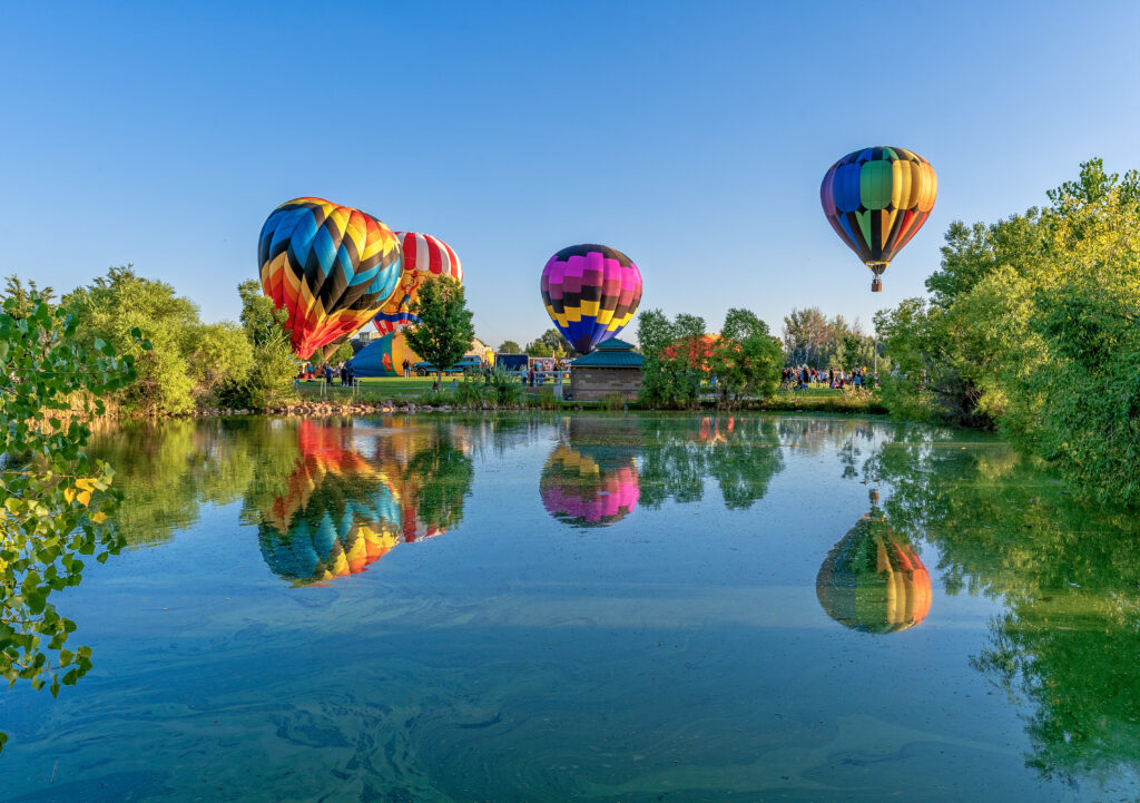Colorful hot air balloons floating above a calm pond, reflecting in the water, surrounded by green trees and a clear blue sky.
