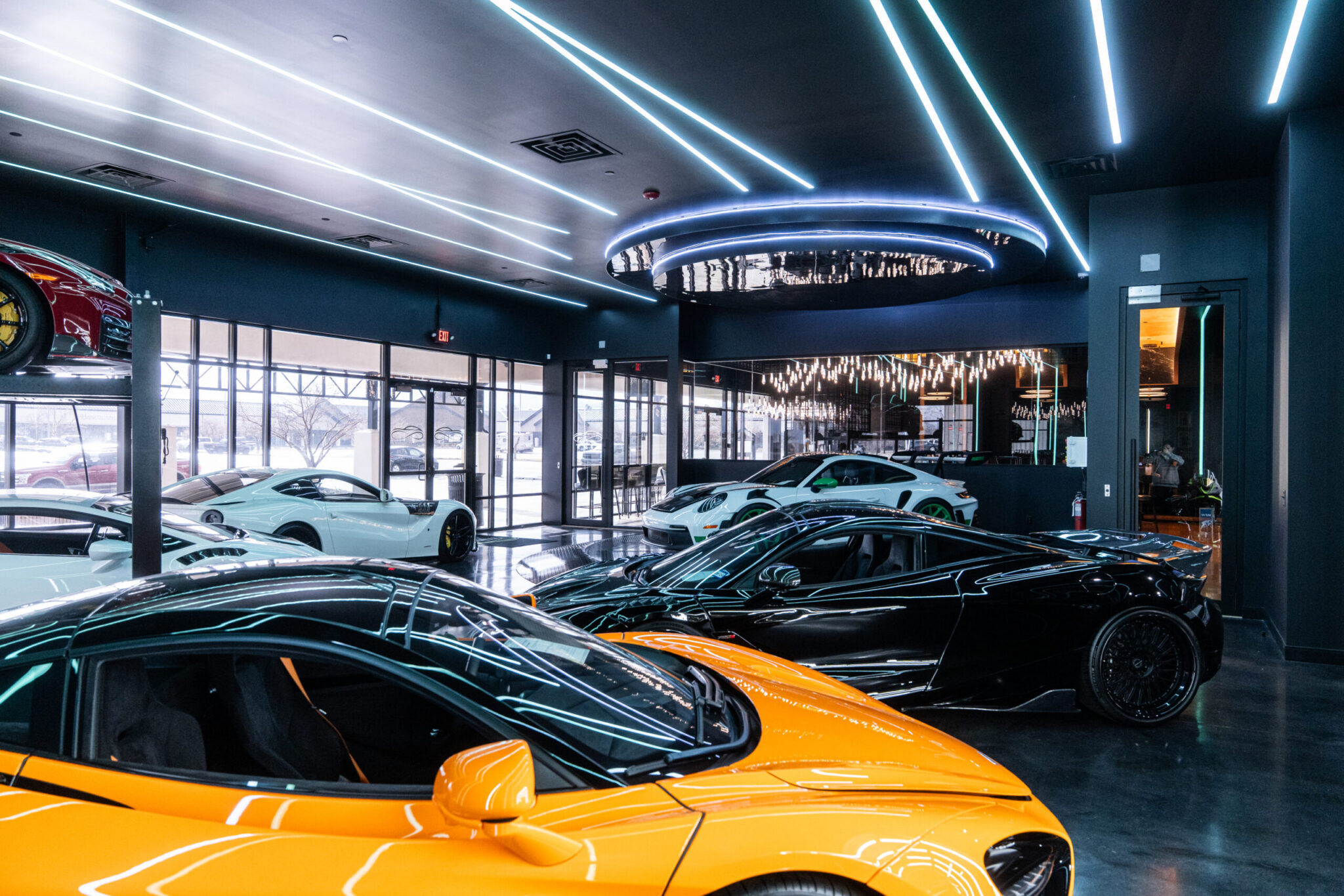 An upscale car showroom featuring multiple luxury sports cars, including an orange one in the foreground and various white and black models in the background. Modern lighting enhances the sleek interior design.