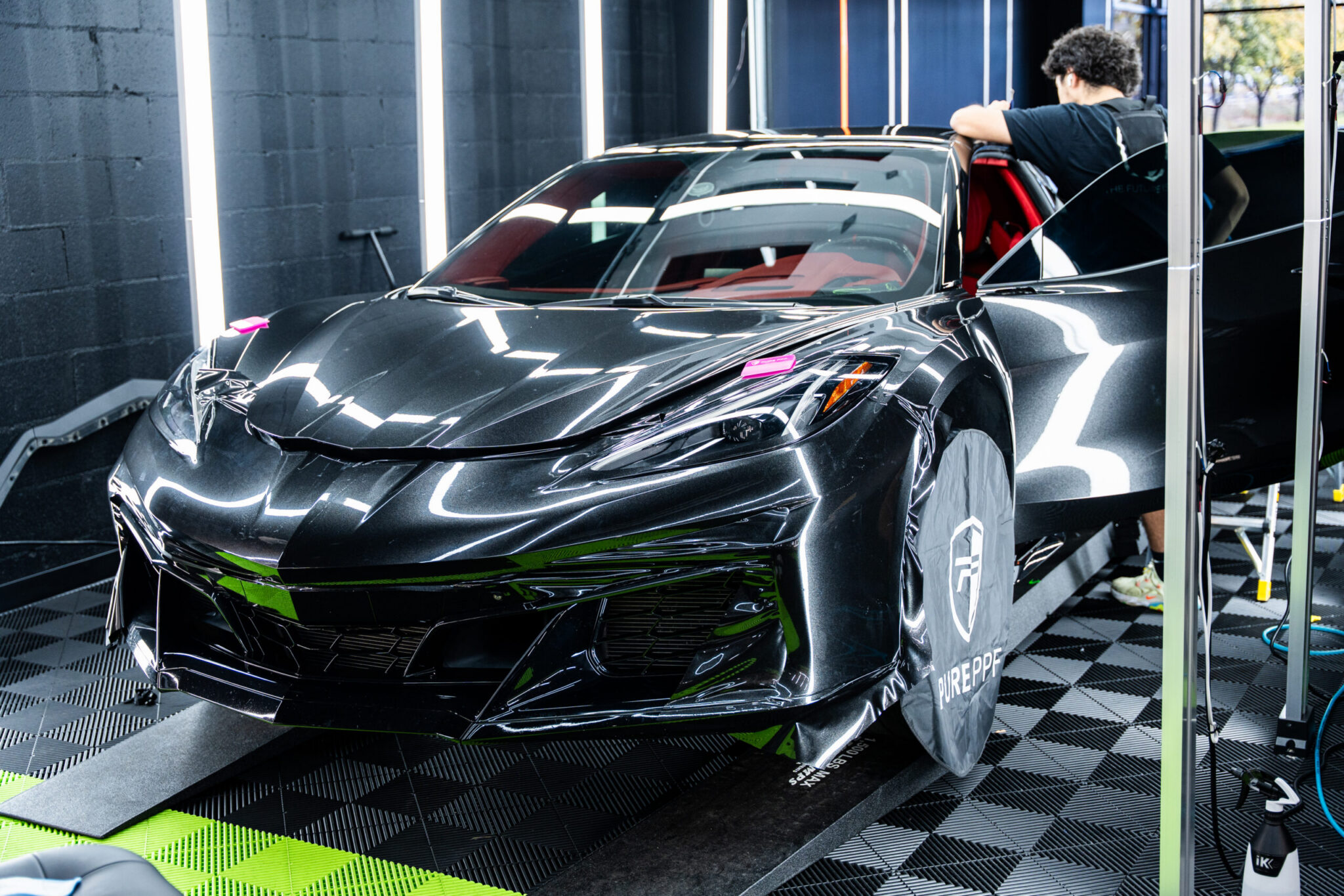 A black sports car with a sleek design, parked in a detailing shop. A person is seen preparing the interior with red accents. Bright lights illuminate the space behind the vehicle.