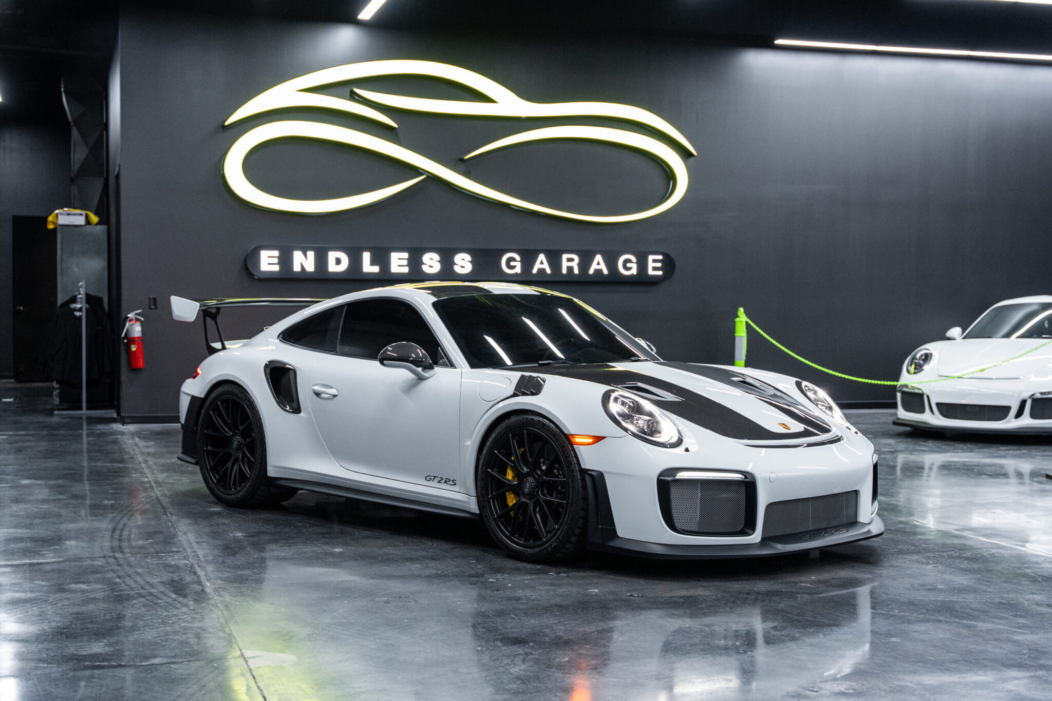 A white sports car with a black racing stripe parked in a modern garage featuring a sleek design. The garage has a sign reading 'ENDLESS GARAGE' on the wall, with another white sports car visible in the background.