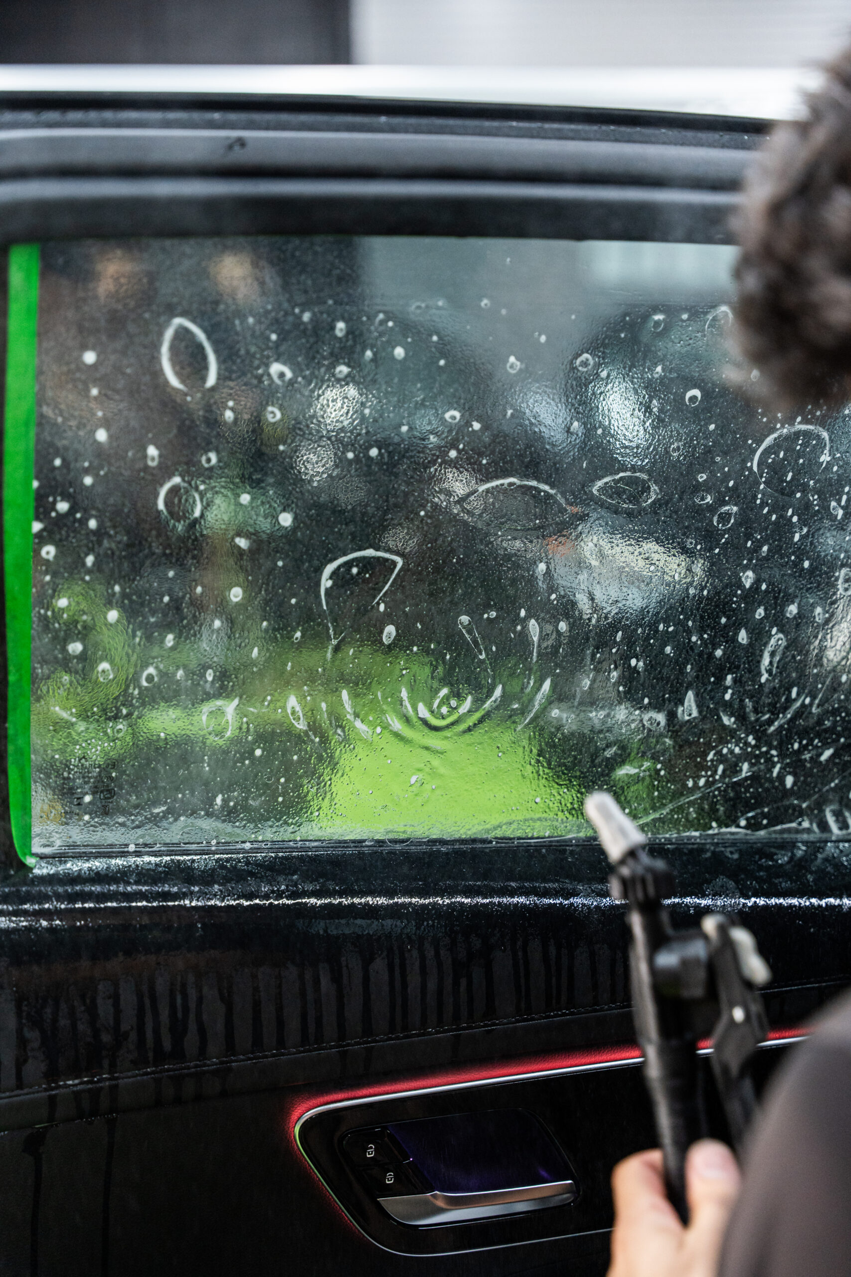 A close-up view of a car window being cleaned. A person is holding a cleaning tool, with bubbles and water droplets visible on the window. The window has a green tape border.