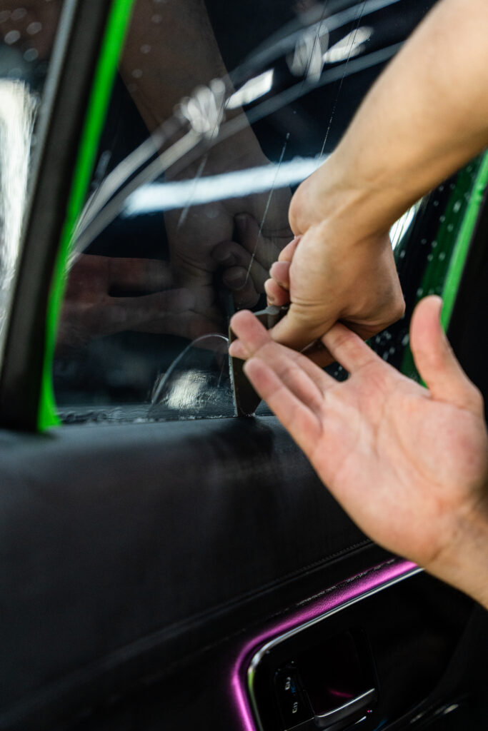A close-up of a person using a tool to repair or remove a piece of window film on a car door, with hands visible and reflections in the glass.