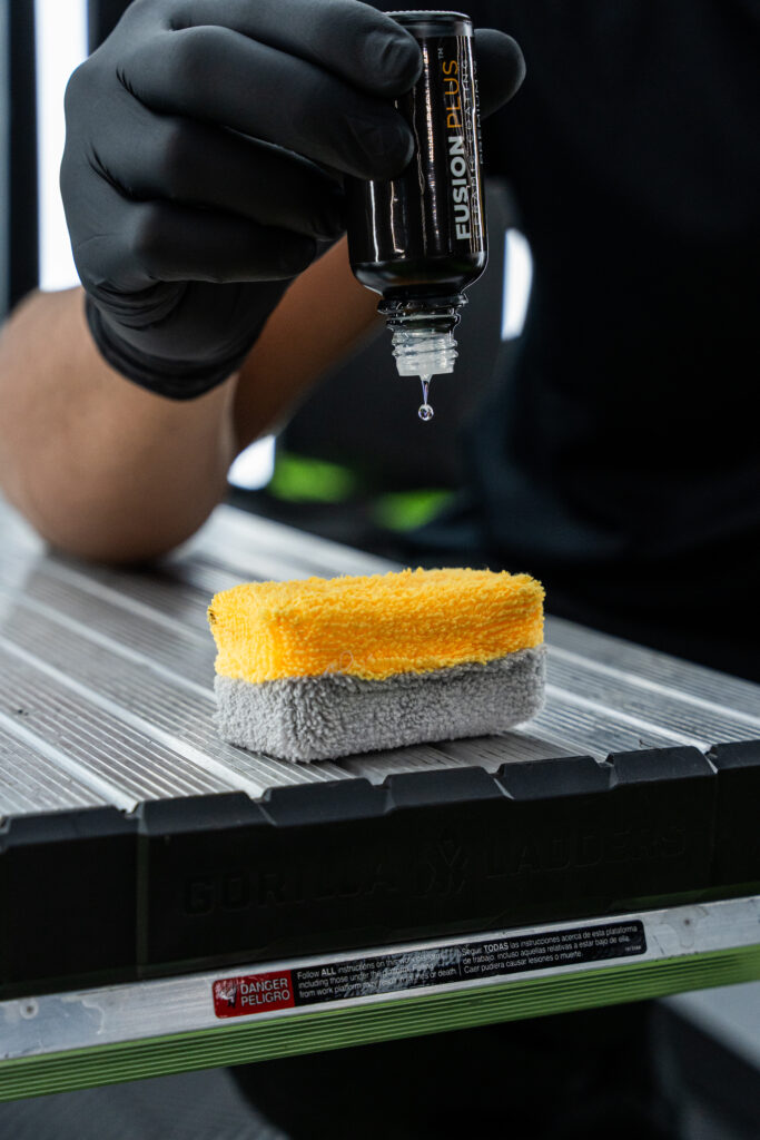 A person wearing black gloves is holding a bottle of car detailing product above a microfiber sponge. The sponge has a yellow top and gray bottom, placed on a metal surface.