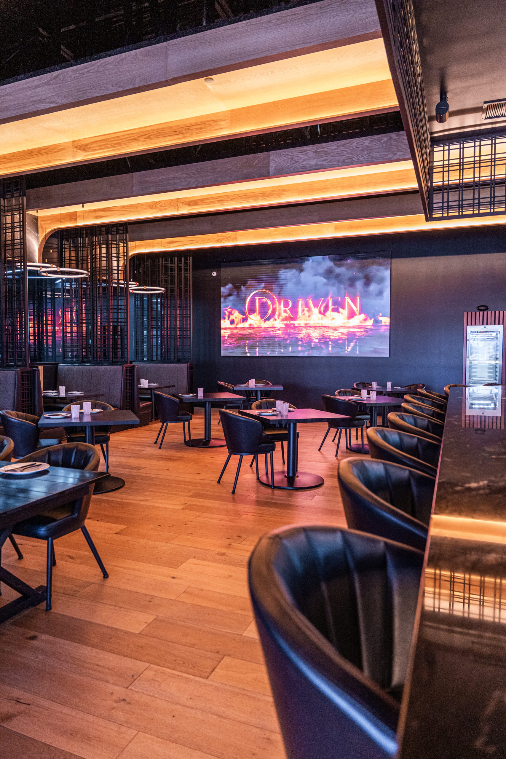 An upscale restaurant interior featuring dark wood floors and modern furniture. Round tables surrounded by black cushioned chairs. A large neon sign reading 'DRIVEN' is illuminated on the wall.