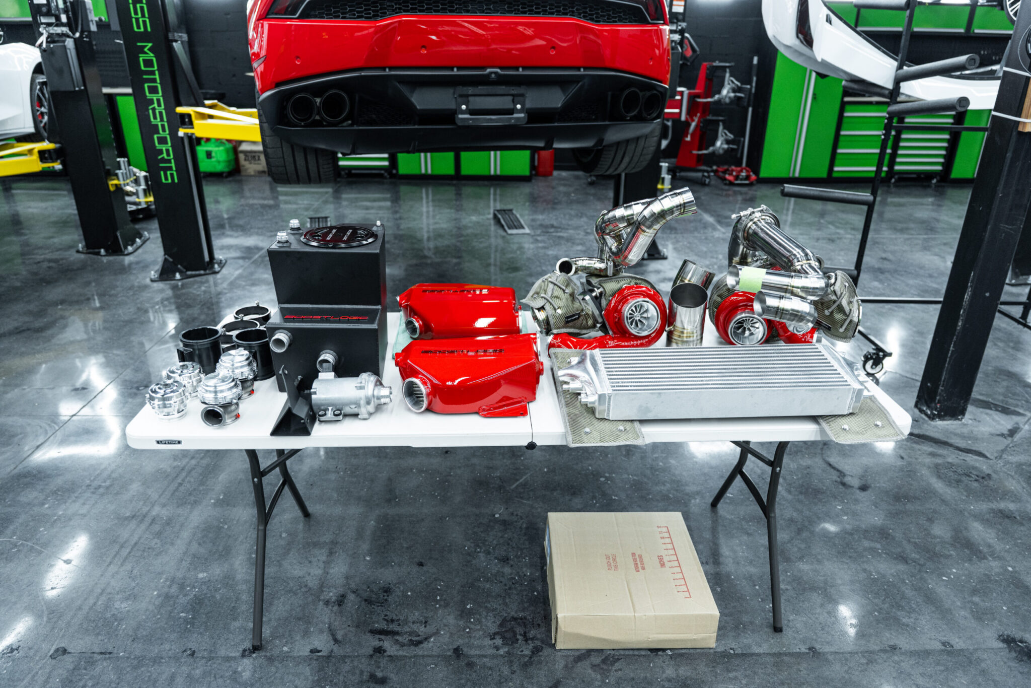 A workbench in a garage displaying various car parts, including red engine components, turbochargers, and an intercooler, with a red sports car partially visible in the background.