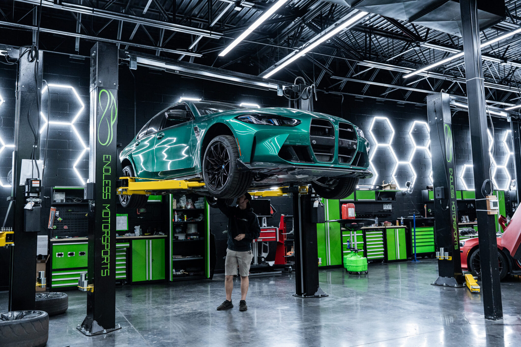 A mechanic working underneath a green sports car raised on a lift in a modern garage. The background features illuminated hexagonal light fixtures and tool storage units.