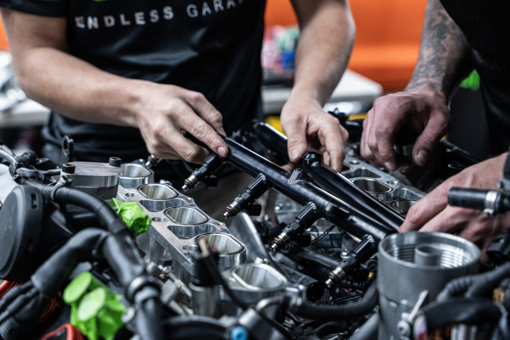 Two mechanics working on an engine in a workshop, holding components while assembling parts. Tools and engine components are visible, with a colorful background hinting at a casual workspace.