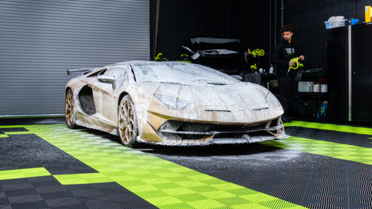 A Lamborghini Aventador covered in white foam, positioned on a green and black patterned floor inside a garage. A person is cleaning the vehicle in the background.