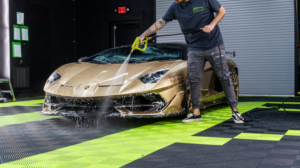 A person using a pressure washer to clean a gold Lamborghini Aventador in a garage. The car is covered in soap, with vibrant green and black flooring visible.